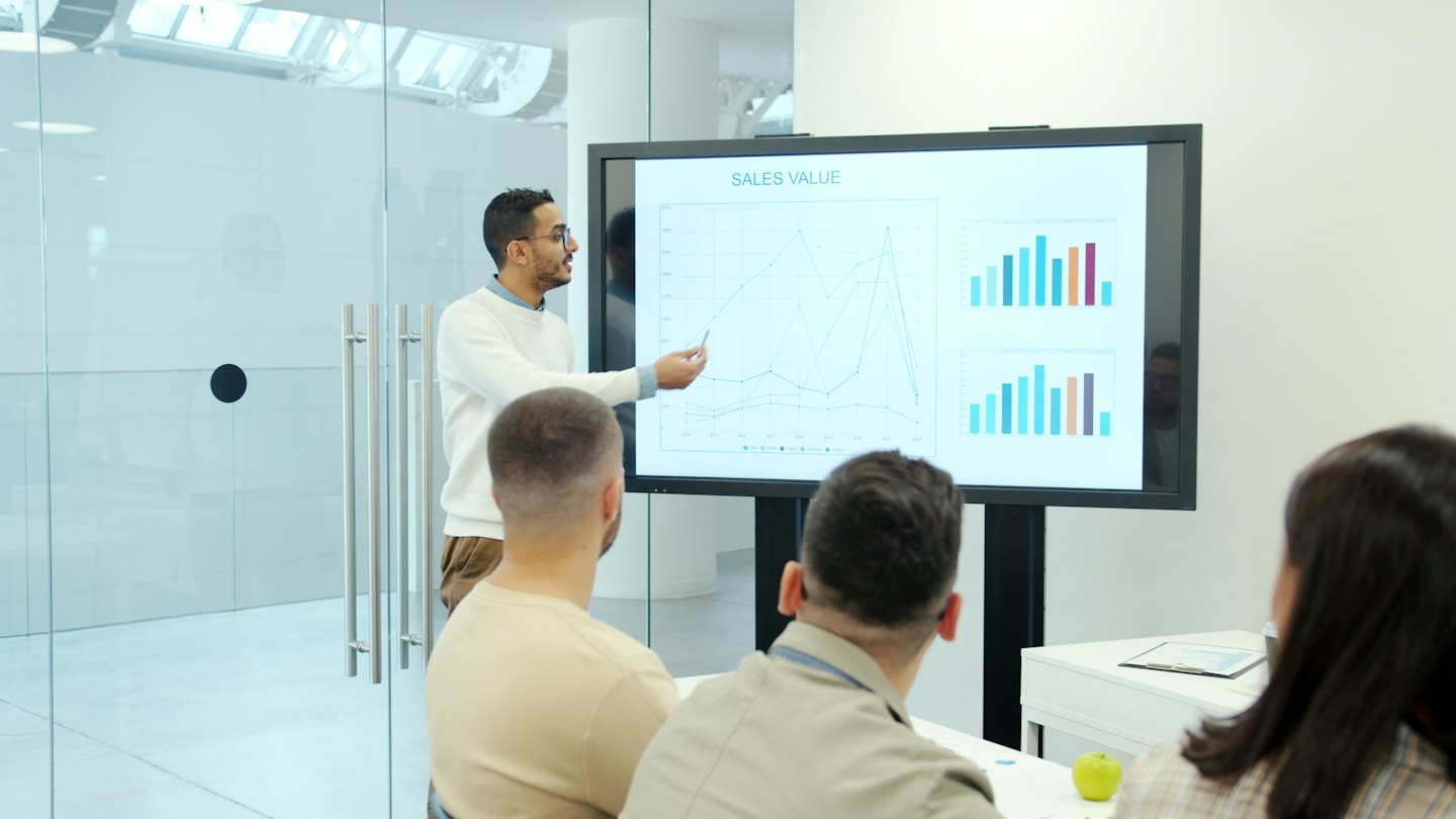 Man presenting sales graphs on screen to colleagues in office.